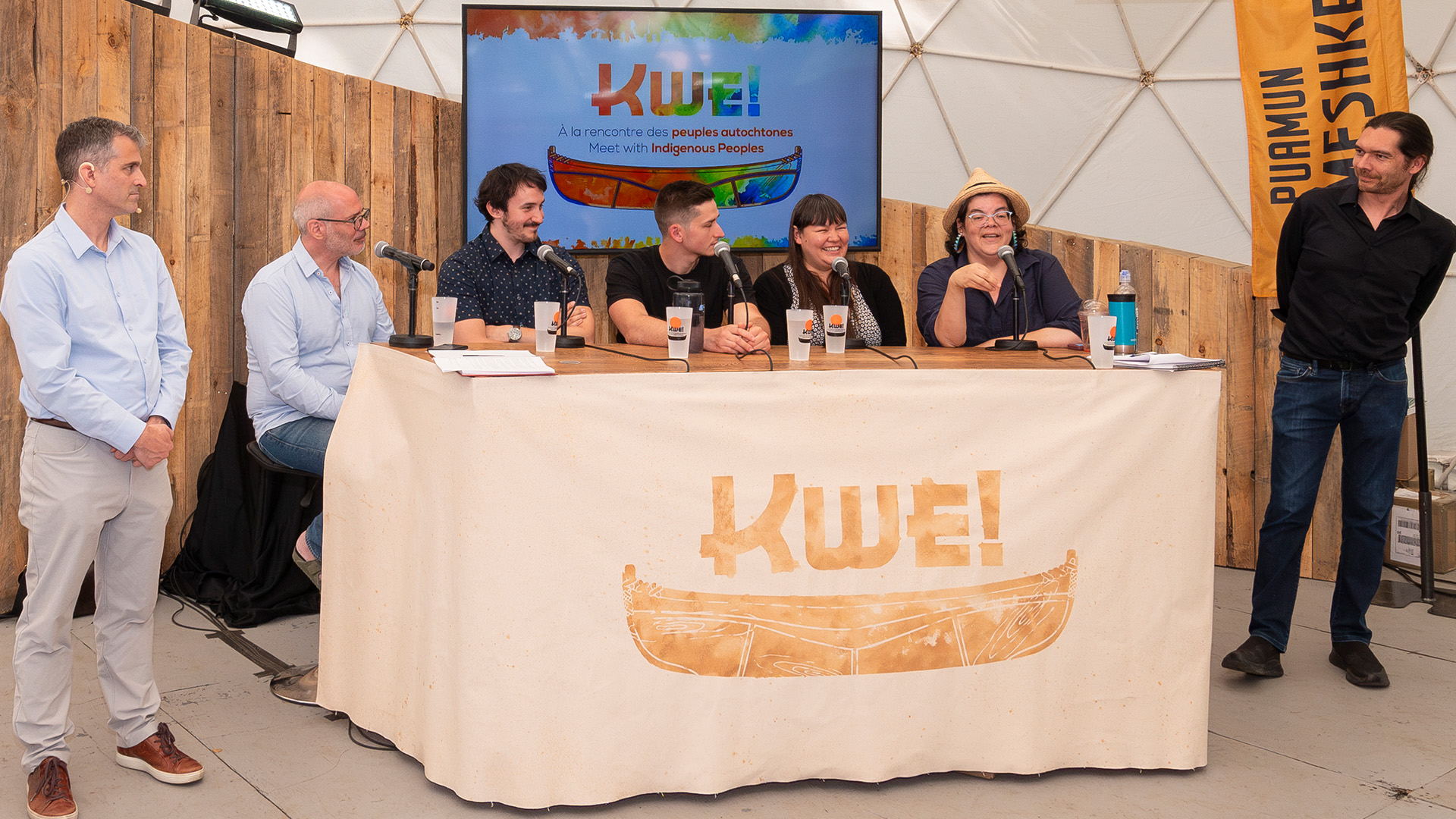 Alex Boissonneault and Alexandre Bacon standing,Jérôme Gill-Couture, Samuel Lapointe-Savard, Melissa Mollen Dupuis,Nahka Bertrand, Mathieu Carli seated at a table covered with a light coloured table cloth with the KWEI letters in gold